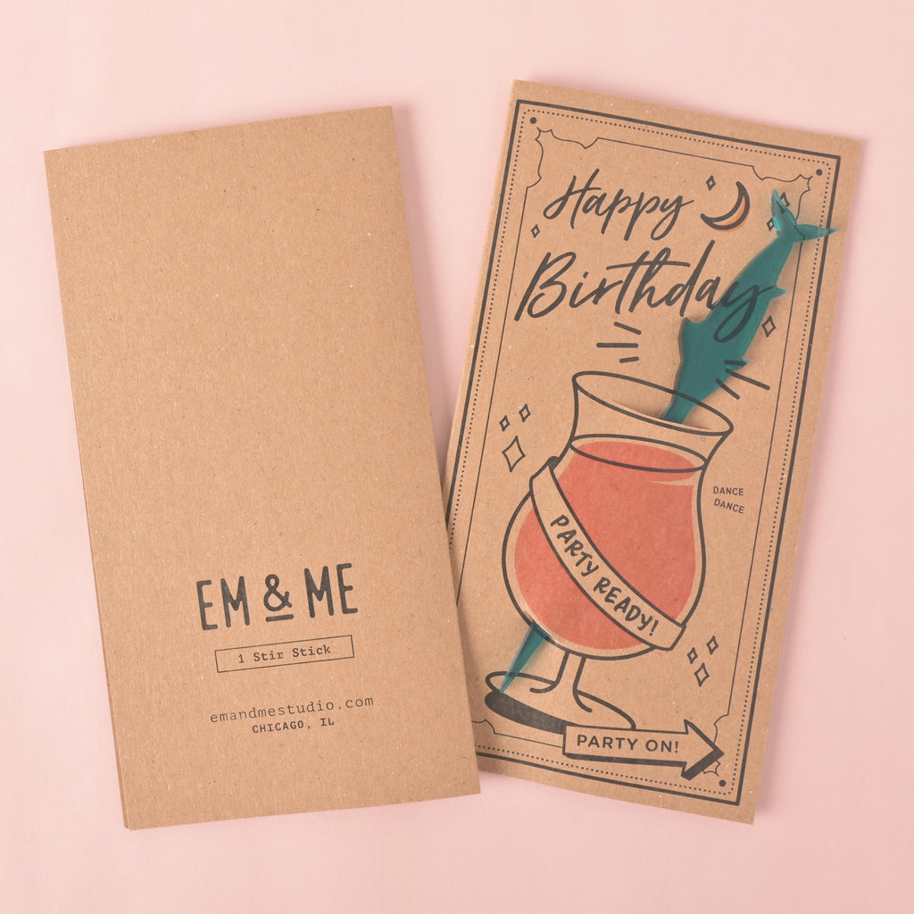 A "Happy Birthday" gift card and stirrer set. The front of the card features a drawing of a cocktail glass with a teal swordfish-shaped stirrer and the words "Party Ready!" The back of the card shows the "EM & ME" logo, "1 Stir Stick," and the company website and location. The items are displayed on a solid pink background.