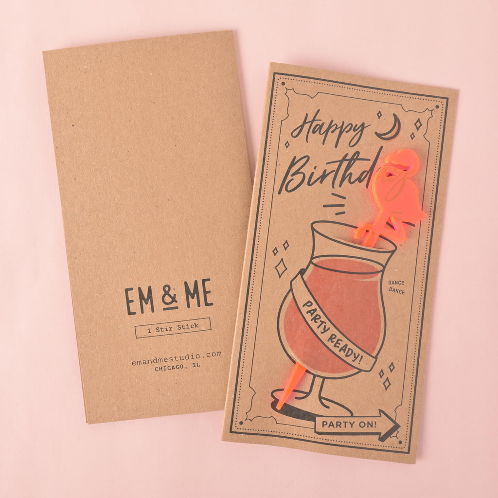 A "Happy Birthday" gift card and stirrer set. The front of the card features a drawing of a cocktail glass with a fluorescent pink flamingo-shaped stirrer and the words "Party Ready!" The back of the card shows the "EM & ME" logo, "1 Stir Stick," and the company website and location. The items are displayed on a solid pink background.