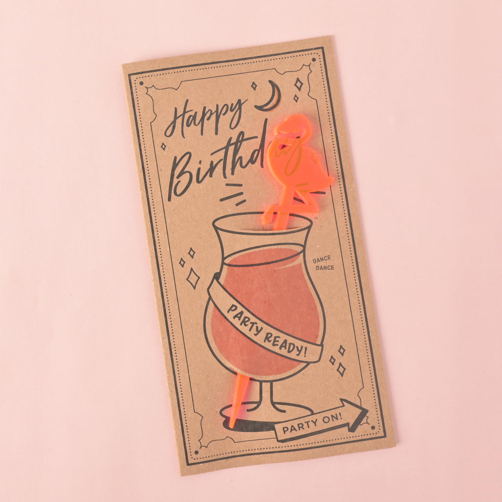 A brown card with a drawing of a cocktail glass and a fluorescent pink flamingo-shaped stirrer attached. The card has "Happy Birthday" and "Party Ready!" printed on it. The item is displayed on a solid pink background.