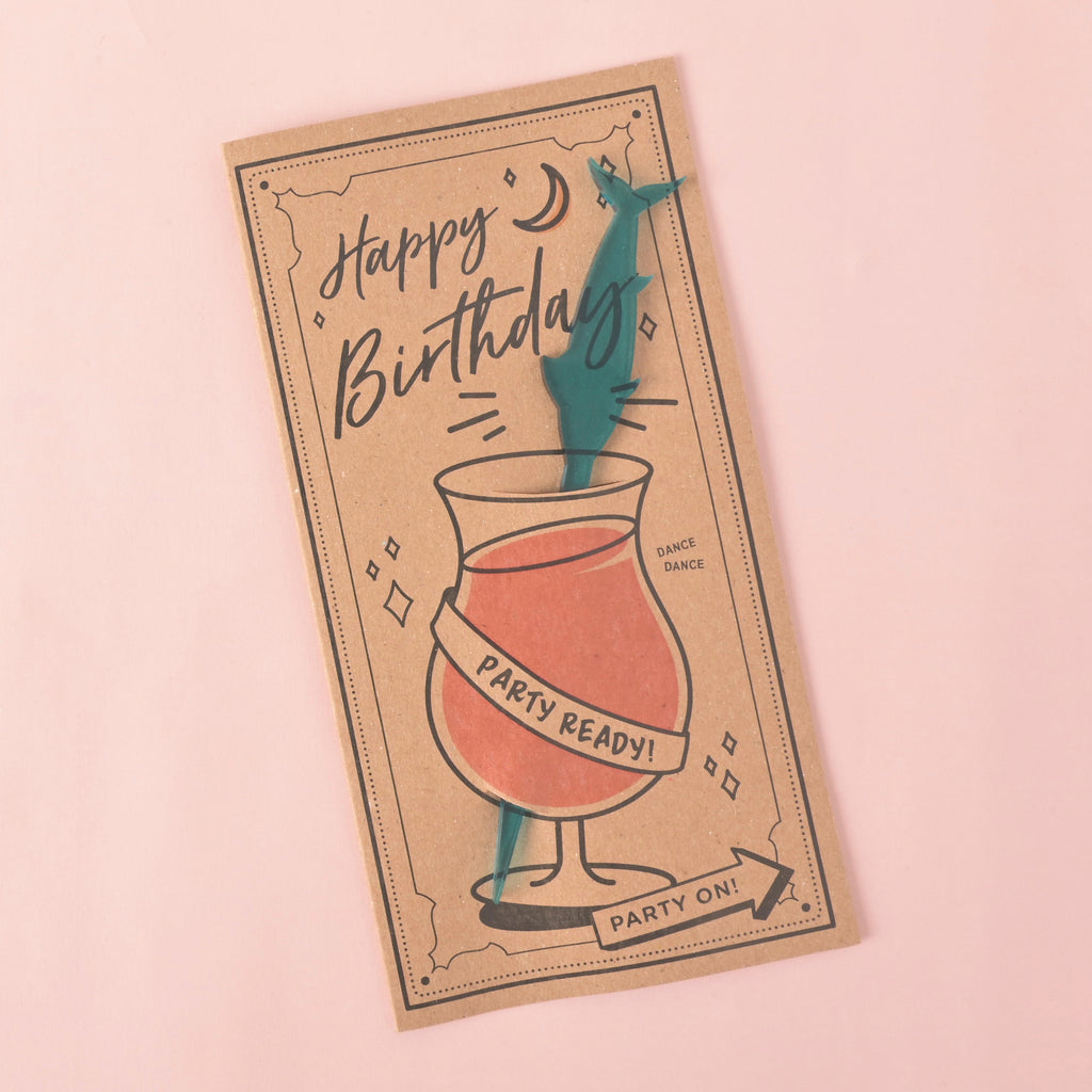 A brown card with a drawing of a cocktail glass and a teal swordfish-shaped stirrer attached. The card has "Happy Birthday" and "Party Ready!" printed on it. The item is displayed on a solid pink background.