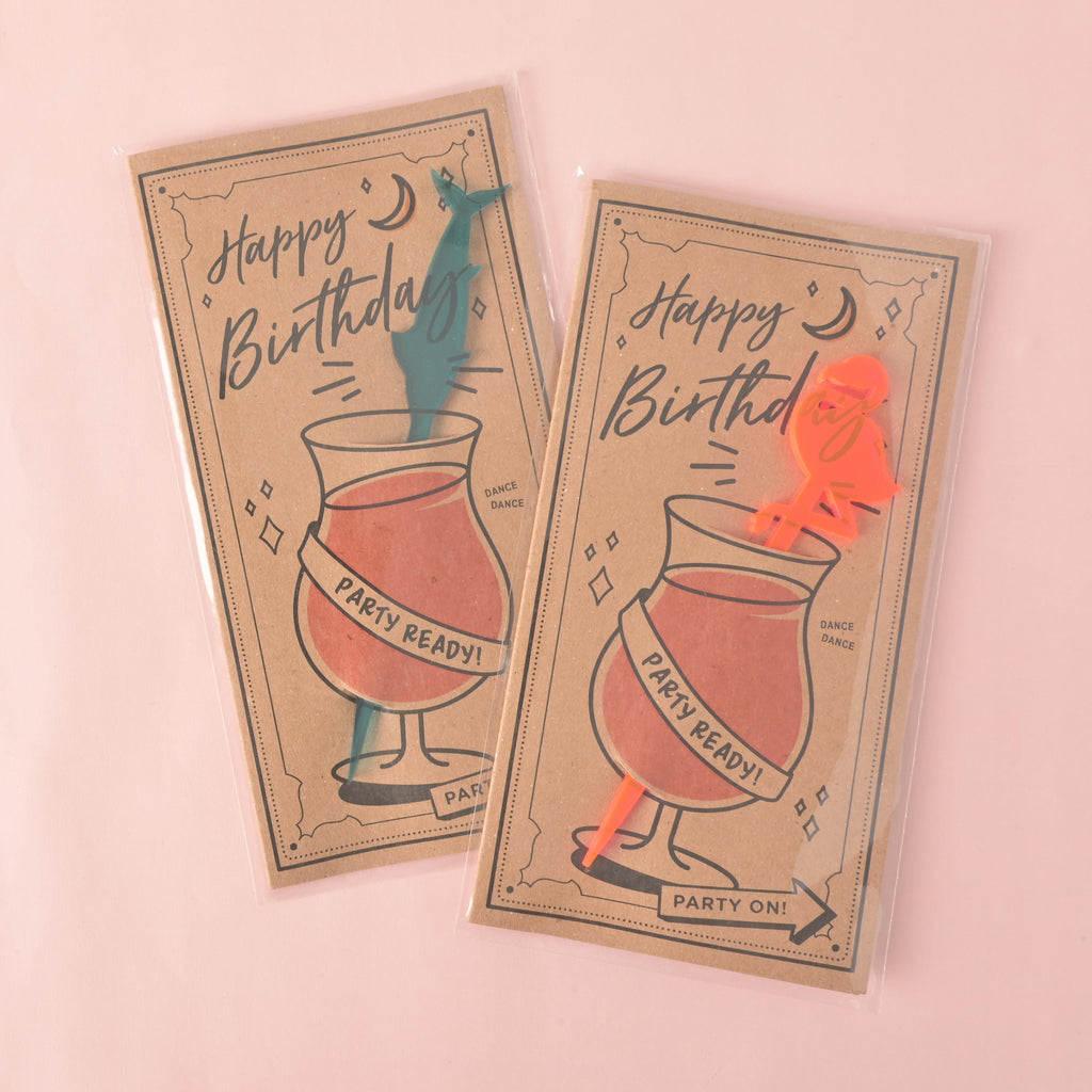 Two "Happy Birthday" gift cards with stirrers sealed in clear plastic. Each card has a drawing of a cocktail glass with the words "Party Ready!" The card on the left has a teal swordfish-shaped stirrer, and the one on the right has a fluorescent pink flamingo-shaped stirrer. The items are displayed on a solid pink background.