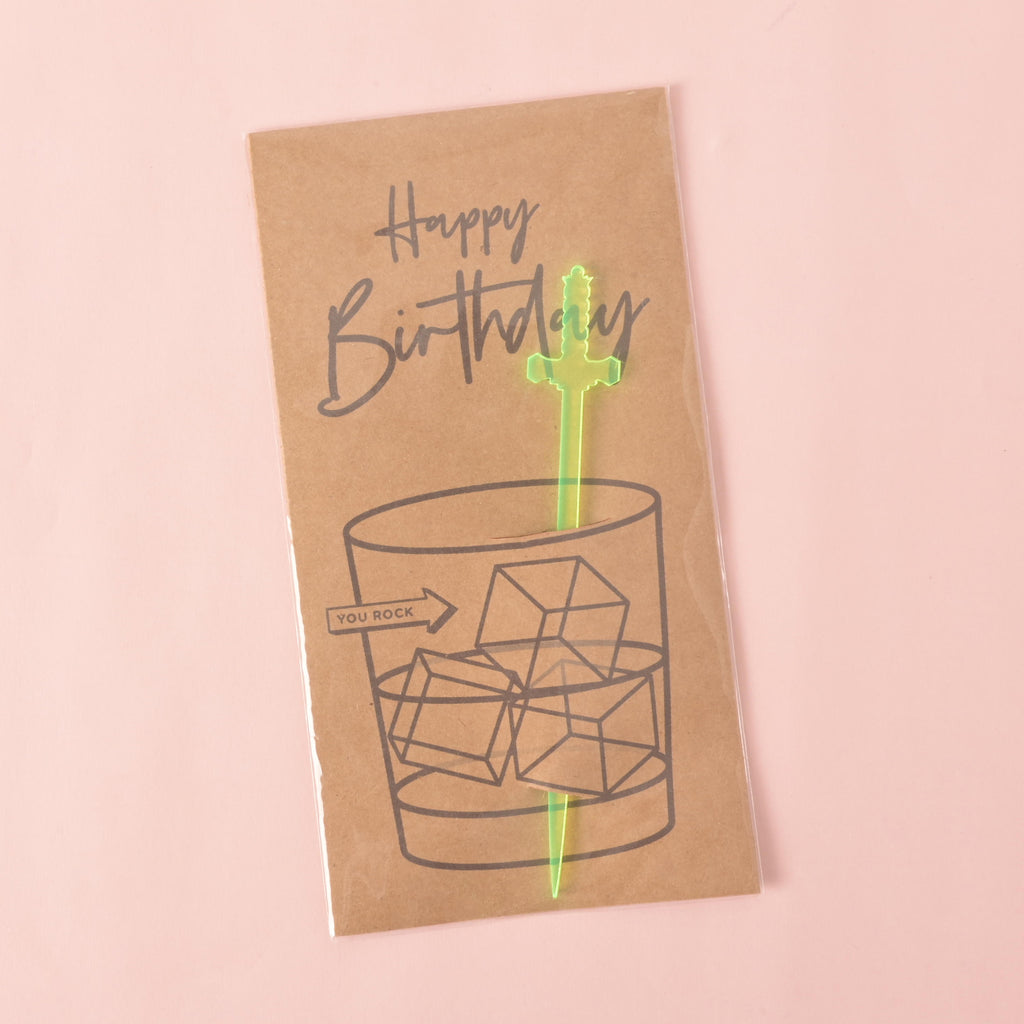 A brown card with a drawing of a glass of ice. It says "Happy Birthday" at the top and "YOU ROCK" on the side. A translucent green sword-shaped cocktail stirrer is attached to the card. The item is displayed on a solid pink background.