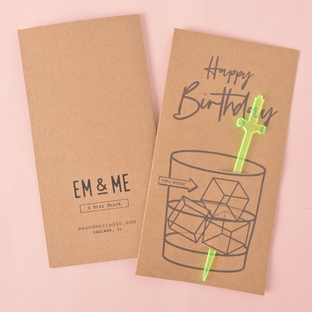 A "Happy Birthday" gift card and stir stick. The front of the card has a drawing of a glass with a translucent green sword-shaped stirrer and the words "You Rock." The back of the card shows the "EM & ME" logo, a bar code, and the company website and location. The items are displayed on a solid pink background.
