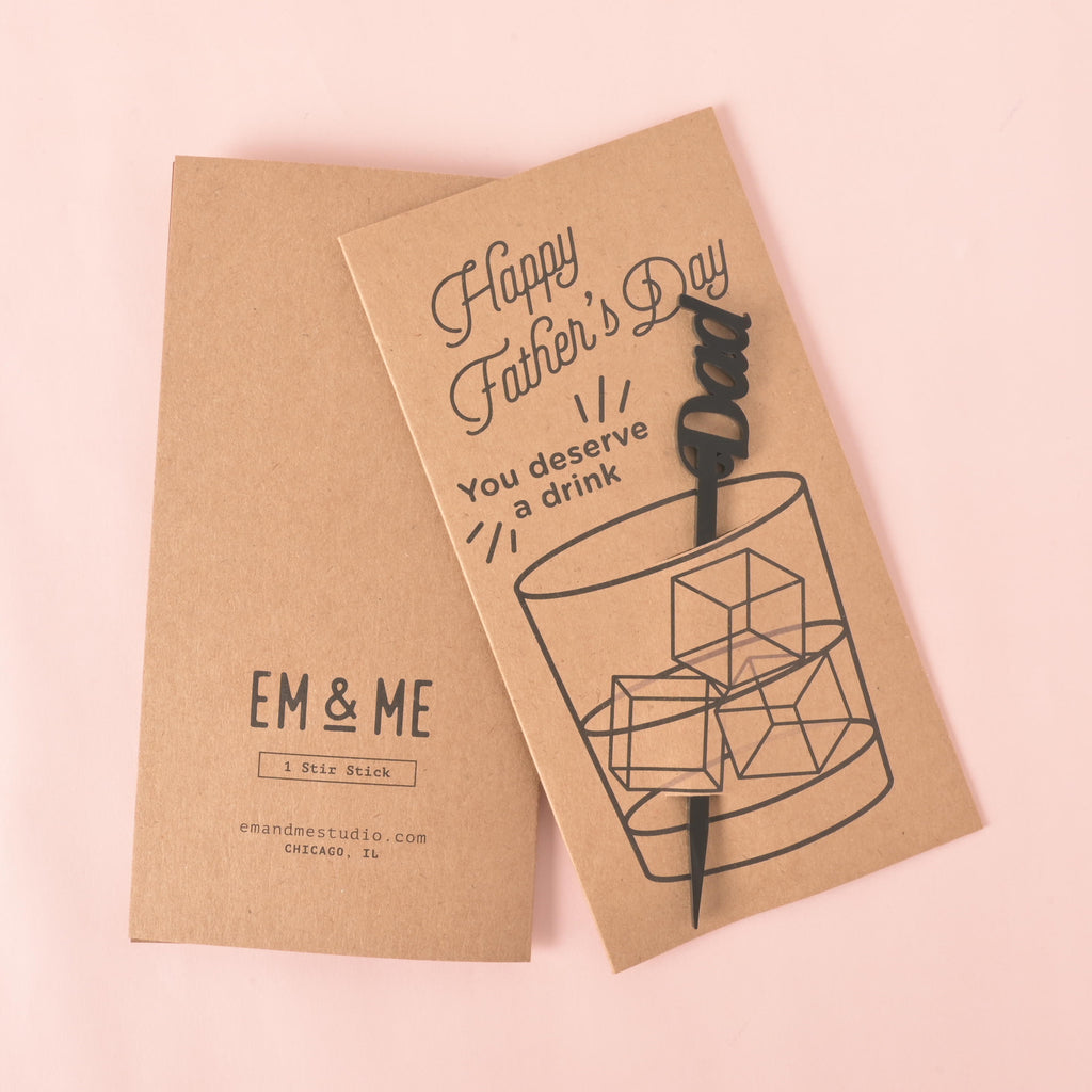 A brown card with a drawing of a glass of ice. It says "Happy Father's Day" at the top and "You deserve a drink" on the side. A black cocktail stirrer with the word "Dad" is attached to the card. The item is displayed on a solid pink background.