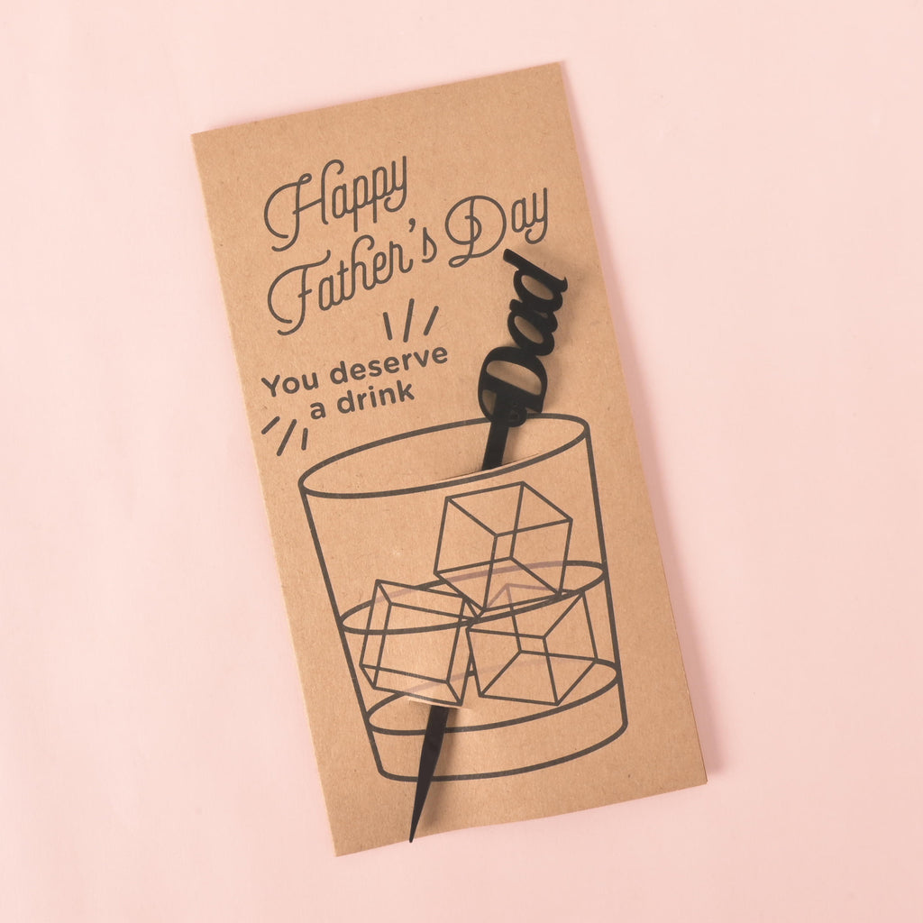 A brown card with a drawing of a glass of ice. It says "Happy Father's Day" at the top and "You deserve a drink" on the side. A black cocktail stirrer with the word "Dad" is attached to the card. The item is displayed on a solid pink background.