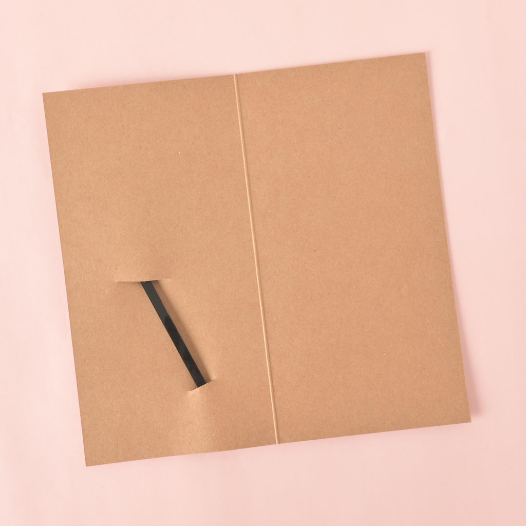 An empty brown gift card, folded in half, with a slit cut into it to hold a stirrer. The back of a black cocktail stirrer is partially visible through the slit. The background is a solid pink color.