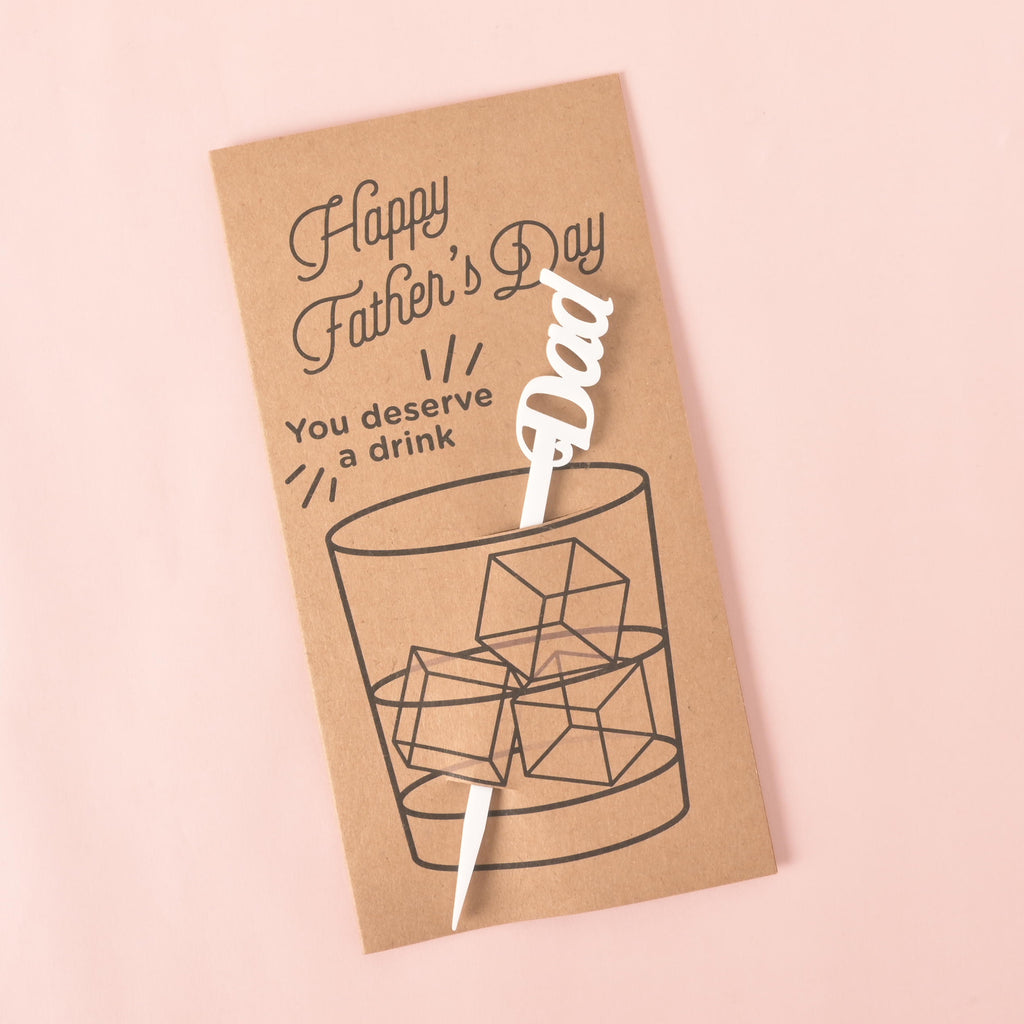A brown card with a drawing of a glass of ice. It says "Happy Father's Day" at the top and "You deserve a drink" on the side. A white cocktail stirrer with the word "Dad" is attached to the card. The item is displayed on a solid pink background.