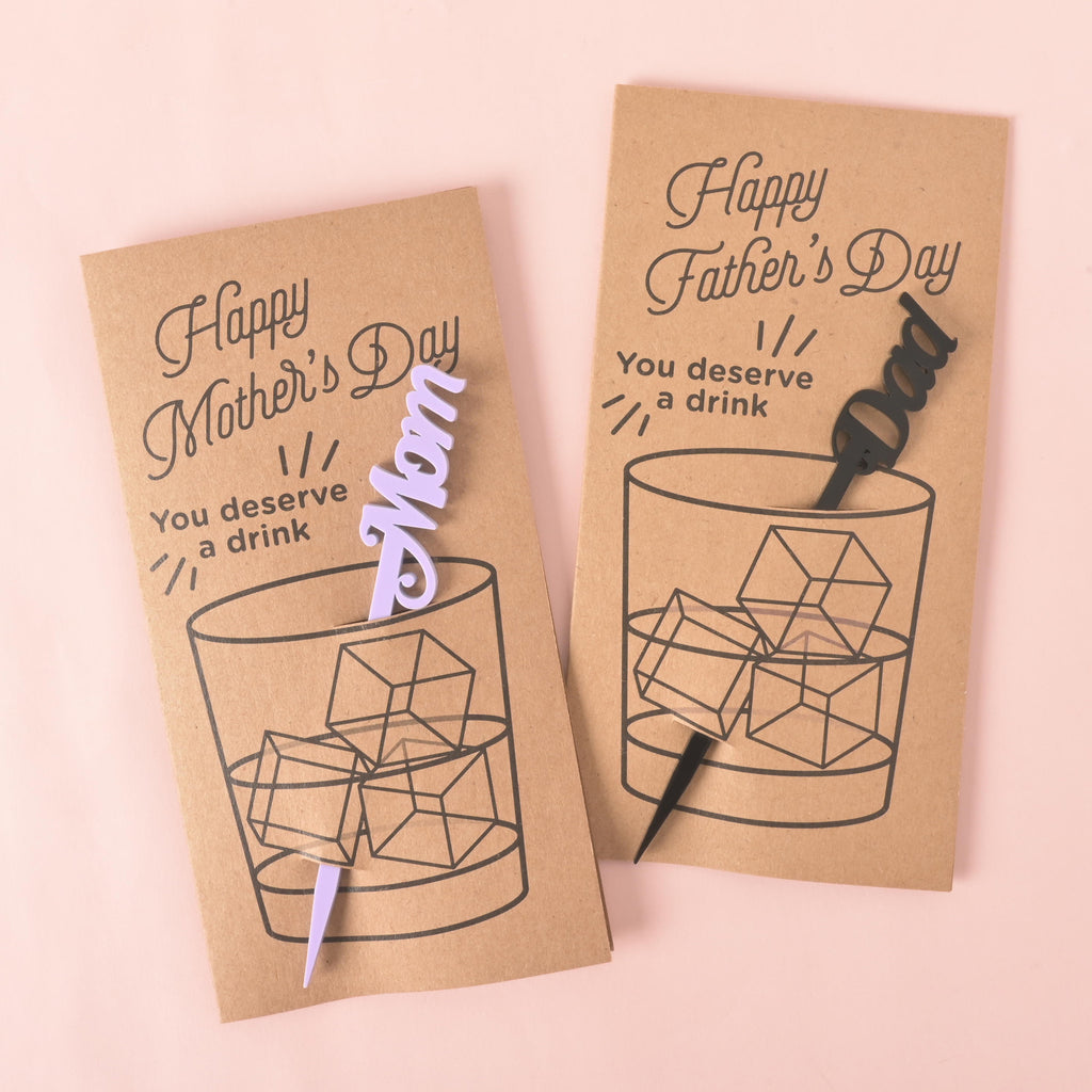 two holiday gift cards with cocktail stirrers. The card on the left says "Happy Mother's Day" and has a light purple "Mom" stirrer. The card on the right says "Happy Father's Day" and has a black "Dad" stirrer. Both cards feature a drawing of a glass with ice and the words "You deserve a drink." The items are displayed on a solid pink background.