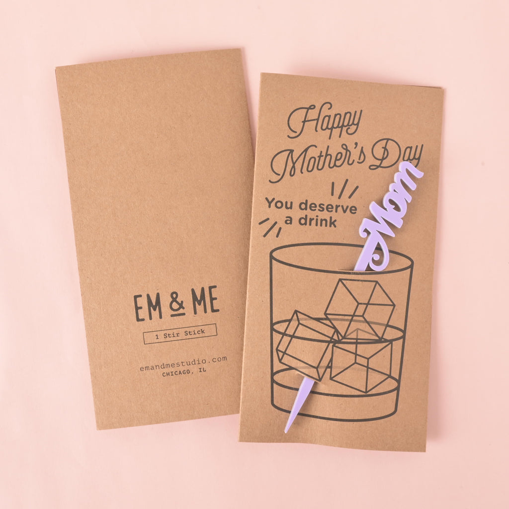 A "Happy Mother's Day" gift card and stirrer set. The front of the card features a drawing of a glass of ice with a purple "Mom" stirrer and the words "You deserve a drink." The back of the card shows the "EM & ME" logo, "1 Stir Stick," and the company website and location. The items are displayed on a solid pink background.