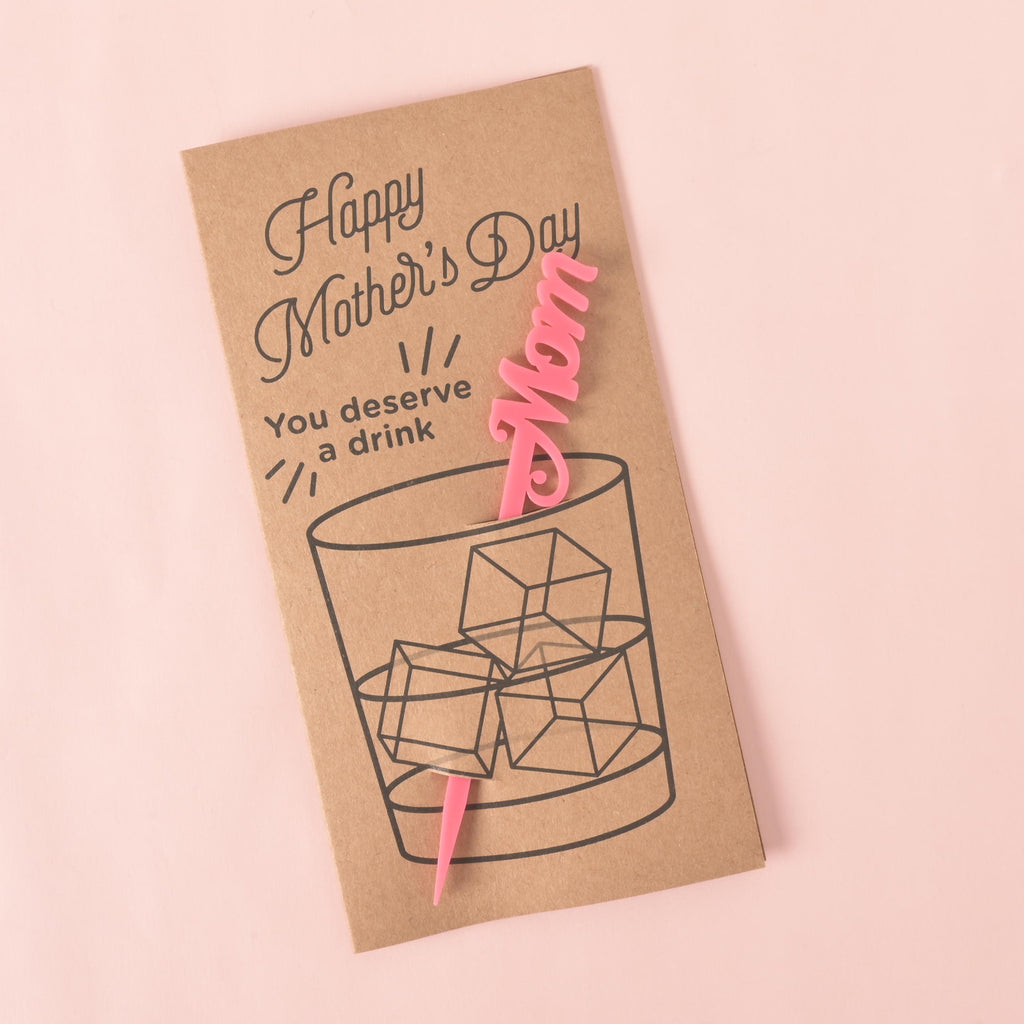 A brown card with a drawing of a glass of ice. It says "Happy Mother's Day" at the top and "You deserve a drink" on the side. A pink cocktail stirrer with the word "Mom" is attached to the card. The item is displayed on a solid pink background.