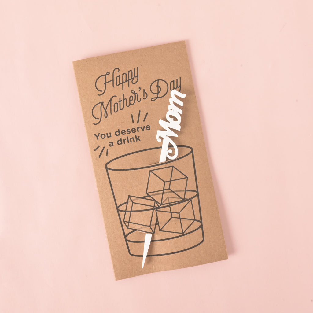 A brown card with a drawing of a glass of ice. It says "Happy Mother's Day" at the top and "You deserve a drink" on the side. A white cocktail stirrer with the word "Mom" is attached to the card. The item is displayed on a solid pink background.