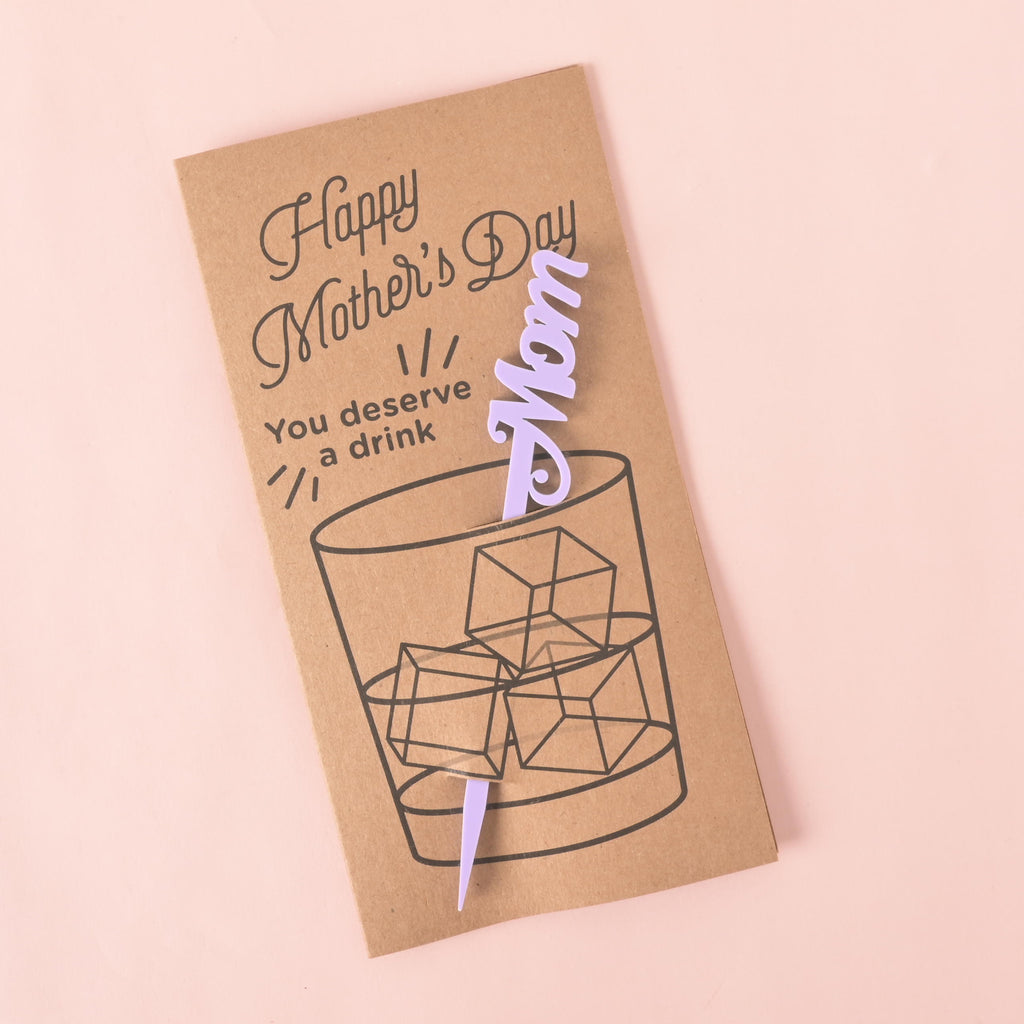 A brown card with a drawing of a glass of ice. It says "Happy Mother's Day" at the top and "You deserve a drink" on the side. A lavender cocktail stirrer with the word "Mom" is attached to the card. The item is displayed on a solid pink background.
