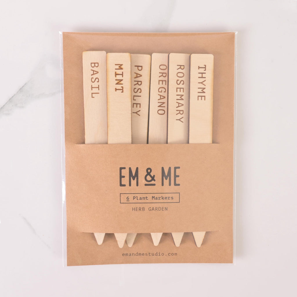 Packaged set of six engraved wooden herb garden markers labeled Basil, Mint, Parsley, Oregano, Rosemary, and Thyme in kraft paper sleeve with EM & ME branding.