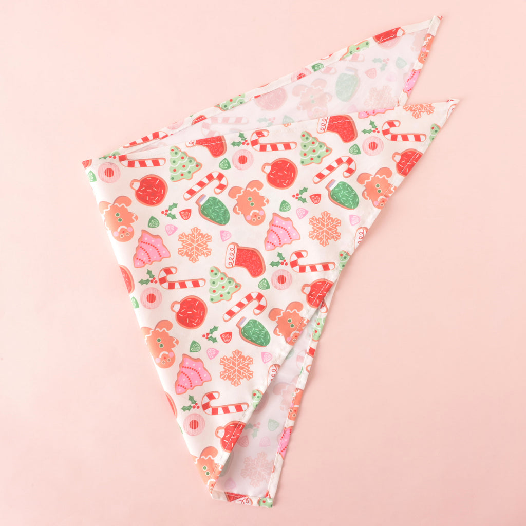 Festive holiday dog bandana with cookie, gingerbread, candy cane, and Christmas tree print on white fabric, folded on a pink background.