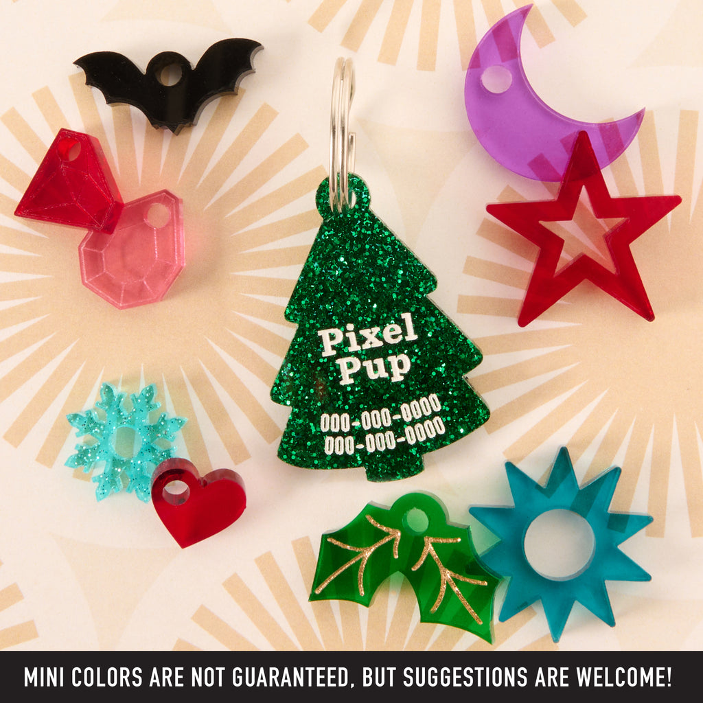 A central green glitter Christmas tree-shaped pet ID tag personalized for 'Pixel Pup.' It is surrounded by assorted small acrylic charms in different colors and shapes: a black bat, red gem, purple crescent moon, red star, blue snowflake, red heart, green holly, and blue sunburst/star shape.