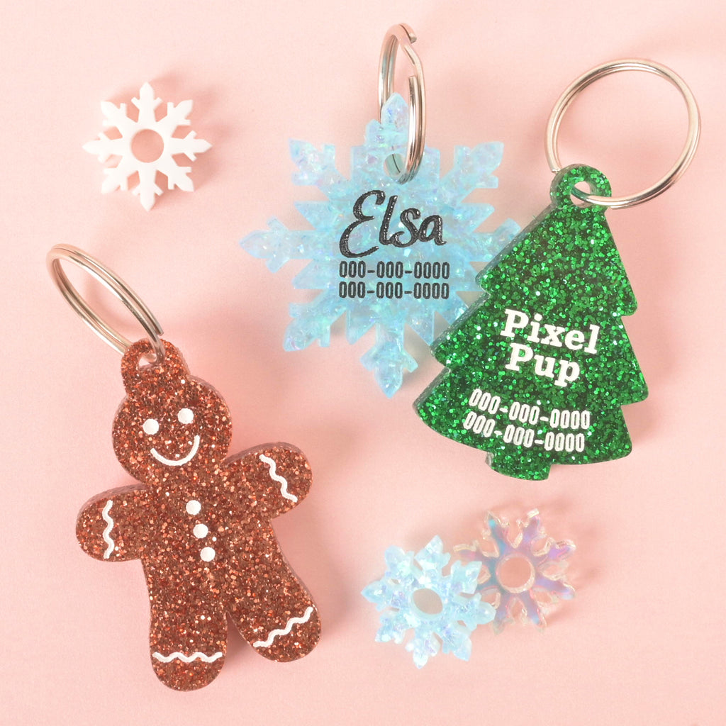 festive pet ID tags including a blue snowflake tag engraved with Elsa, a green glitter Christmas tree tag engraved with Pixel Pup, and a brown glitter gingerbread man tag, displayed with mini snowflake charms on a pink background.