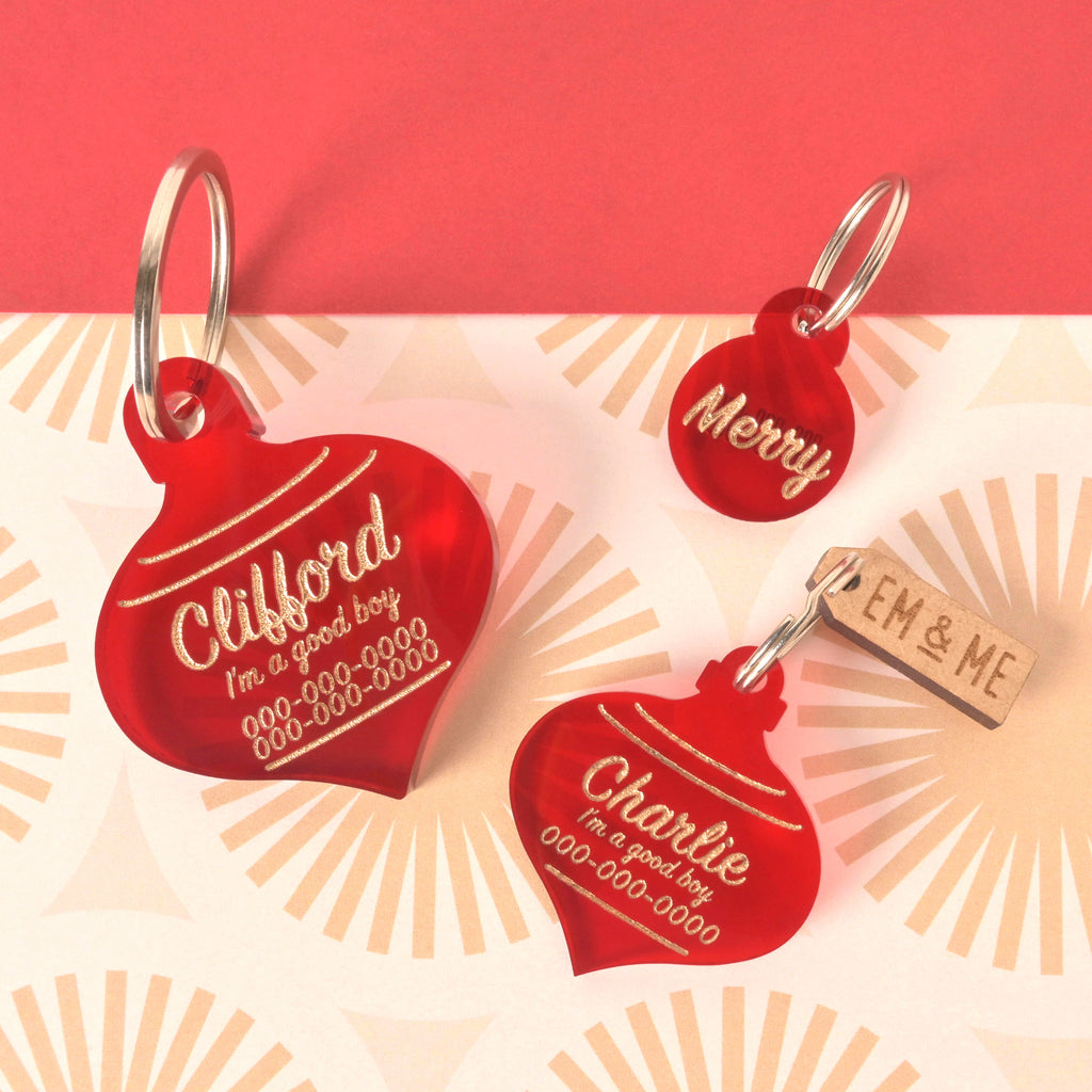 Three translucent red acrylic pet ID tags shaped like Christmas ornaments, featuring gold engraved text. The large tag is personalized for 'Clifford,' a medium tag is personalized for 'Charlie,' and a small round tag reads 'Merry.' A small wooden EM & ME tag is also present. They are displayed on a retro sunburst patterned background.