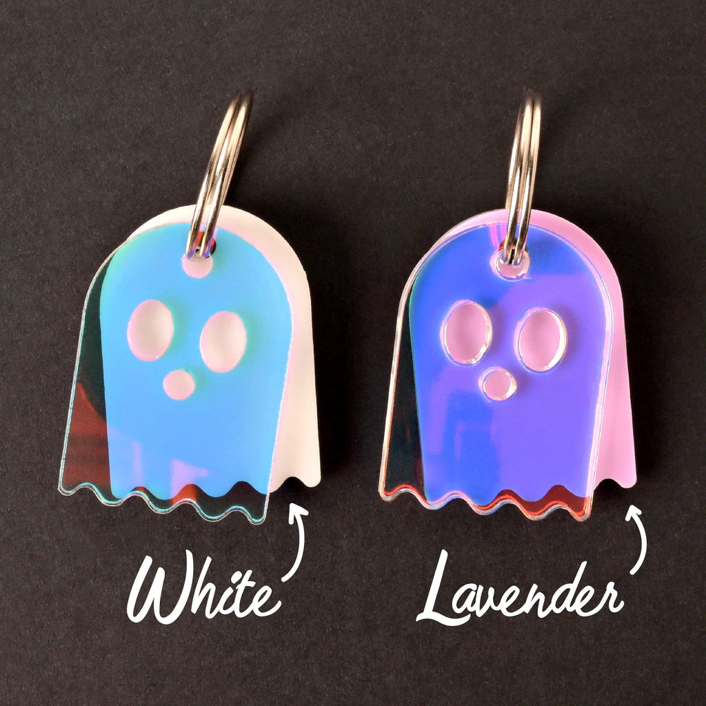A comparison shot of two ghost-shaped pet ID tags on a black background. Both tags are attached to silver keyrings and have a shiny, iridescent, holographic finish. The ghost on the left is labeled "White" and shows a blue and pink sheen. The ghost on the right is labeled "Lavender" and has a prominent purple and pink shine.