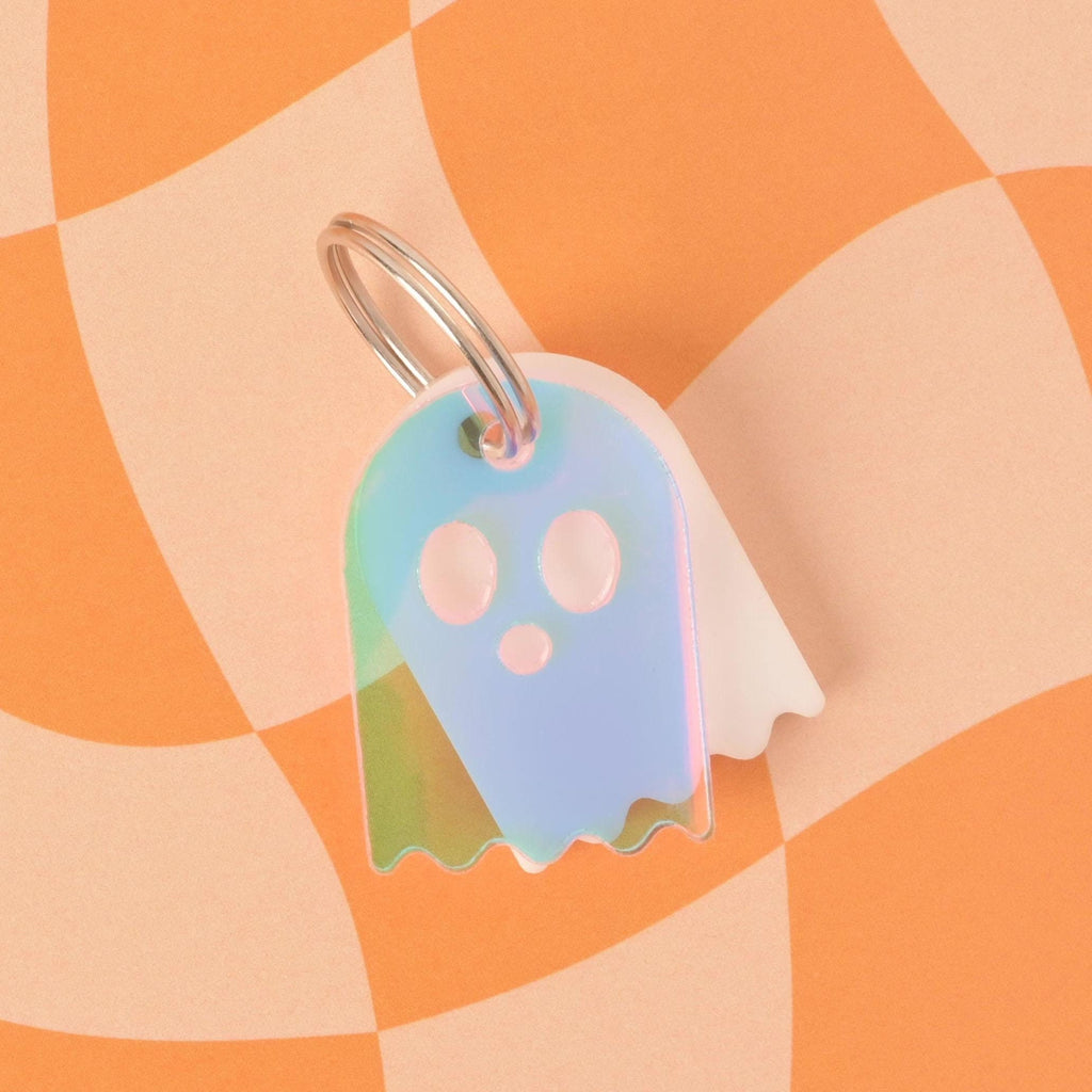 A ghost-shaped pet ID tag with a wavy bottom. The front of the tag is a translucent iridescent material that reflects blue, green, and pink light. A white ghost tag is visible behind it. Both are attached to a silver keyring and are displayed on a checkered orange and beige background.