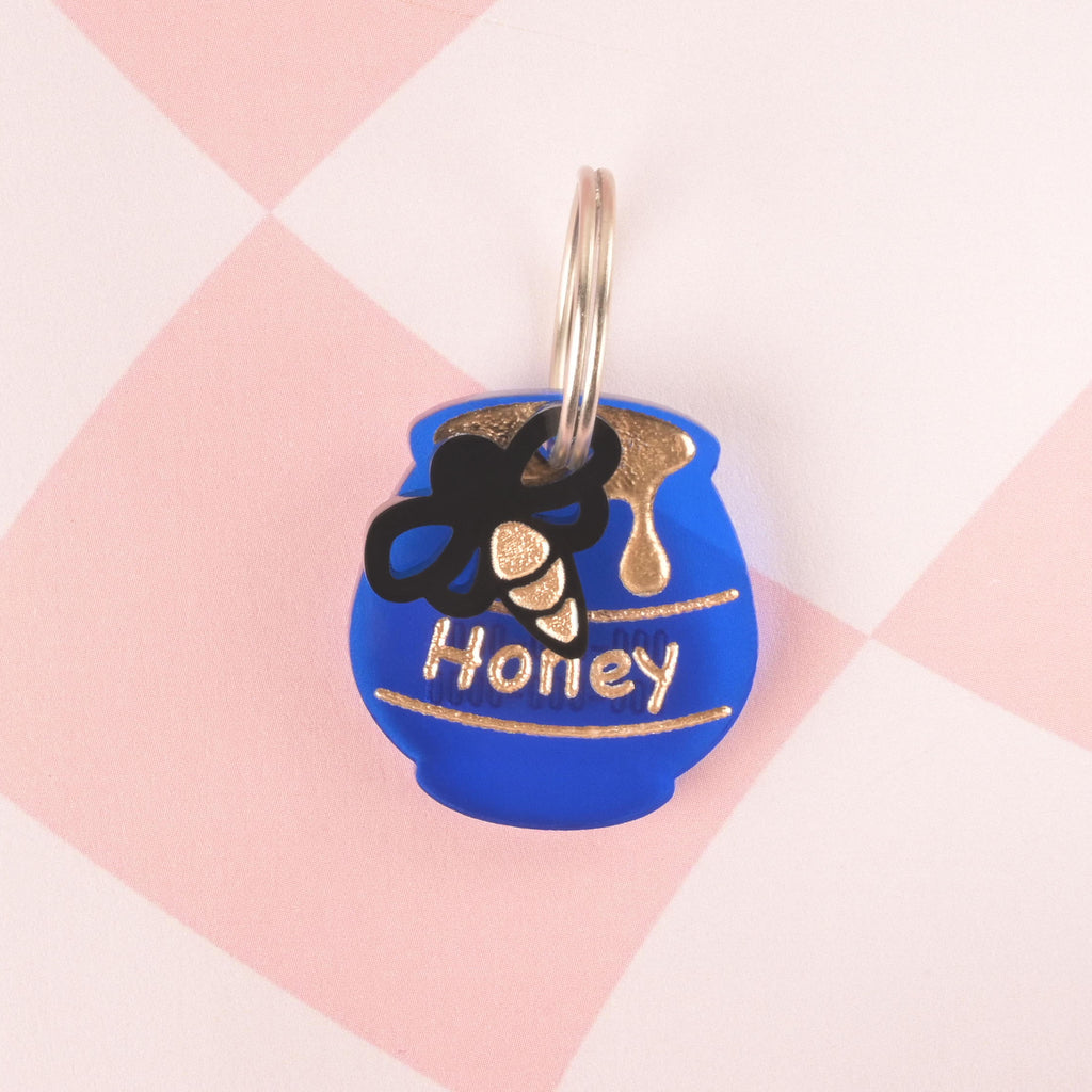 A personalized, blue honey pot-shaped pet tag that says "Honey" in gold letters. The pot has dripping honey details and a small, black bee charm attached. Both are secured with a silver ring, perfect for a collar, and are resting on a pink and white checkered background.