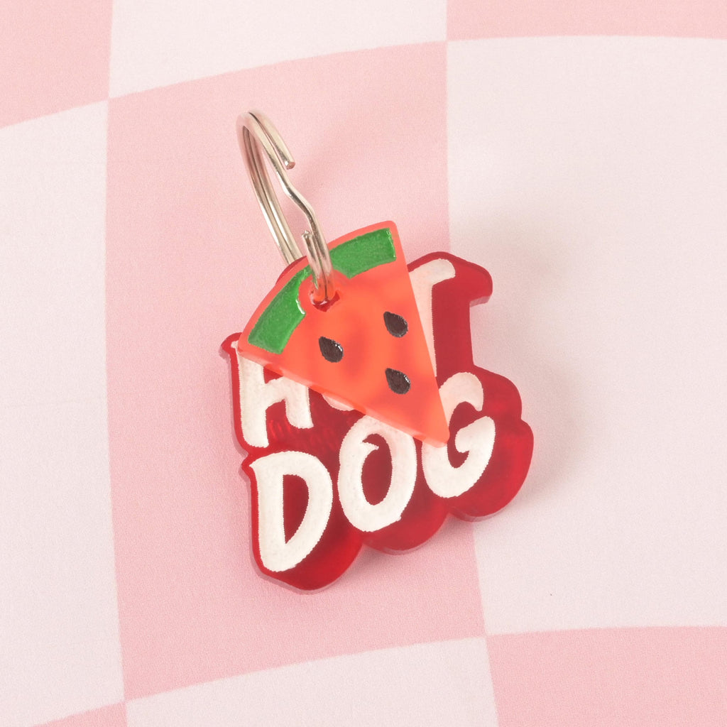A custom, red, translucent pet ID tag shaped like a blob with a white engraving that says "HOT DOG." A small, pink and green watermelon slice charm with black seeds is attached to the shiny silver keyring, sitting in front of the main tag. The tags are displayed on a pink and white checkered background.