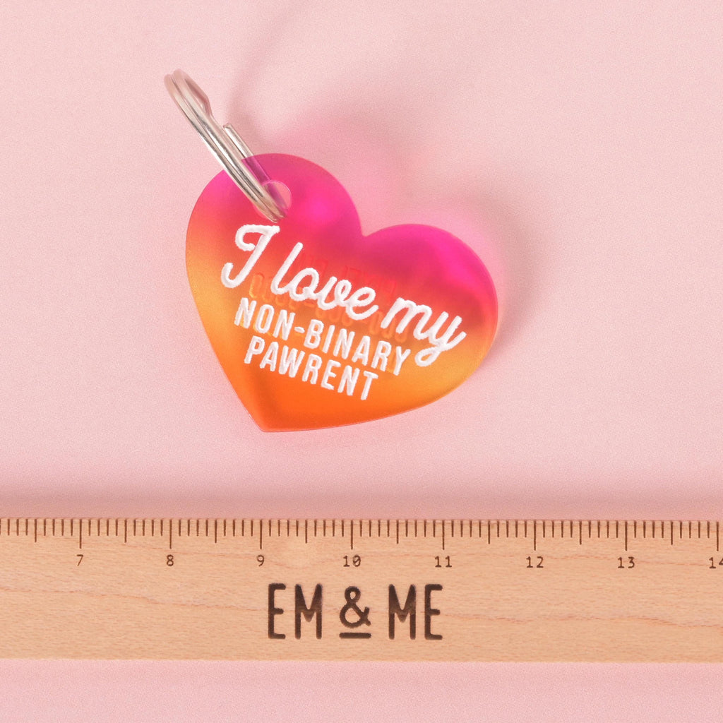 Rainbow gradient heart-shaped pet ID tag with white text that reads “I love my non-binary pawrent,” shown next to a ruler marked EM & ME for size reference. LGBTQ+ pride accessory for dogs and cats.