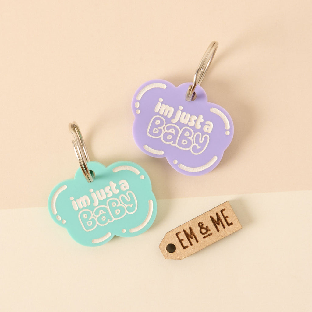 A set of two matching cloud-shaped pet tags that say "i'm just a Baby" in white letters. One tag is light purple, and the other is light blue. A small wooden tag that says "EM & ME" is also shown. All items are attached to a silver ring for a collar and are displayed on a background of light pink and beige.