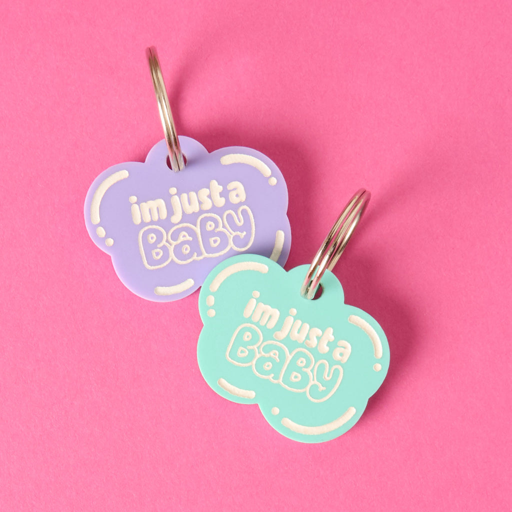 A pair of matching cloud-shaped pet tags that say "i'm just a Baby" in white letters. One tag is light purple and the other is aqua. Both tags are attached to a silver ring, perfect for a collar, and are displayed on a vibrant solid pink background.
