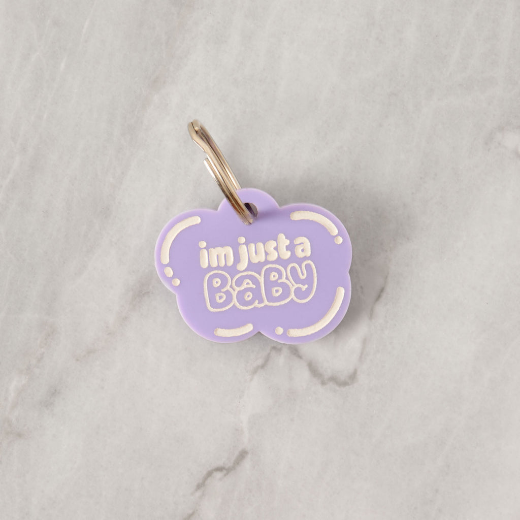 A personalized Lavender cloud-shaped pet tag that says "i'm just a Baby" in white letters. The tag is attached to a silver ring, perfect for a collar, and is displayed on a white marble surface.