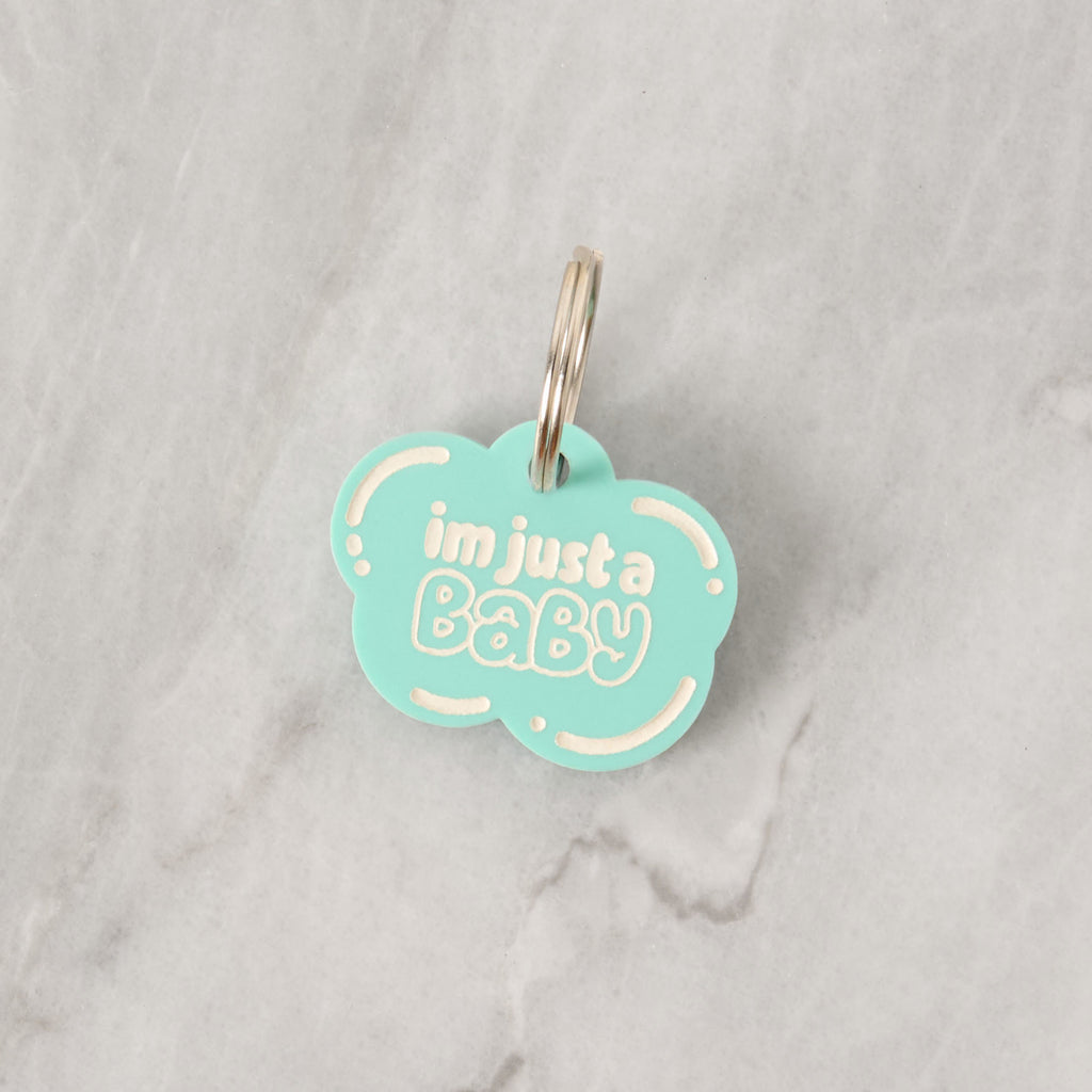 A personalized aqua cloud-shaped pet tag that says "i'm just a Baby" in white letters. The tag is attached to a silver ring, perfect for a collar, and is displayed on a white marble surface.