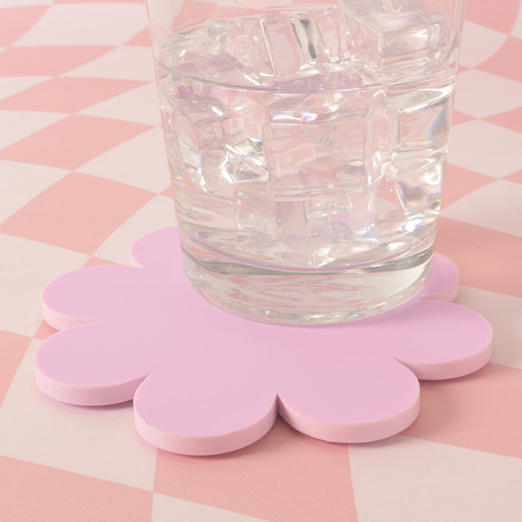 A clear glass filled with ice and a drink, resting on a lavender flower-shaped coaster. The background is a pink and white checkered pattern.