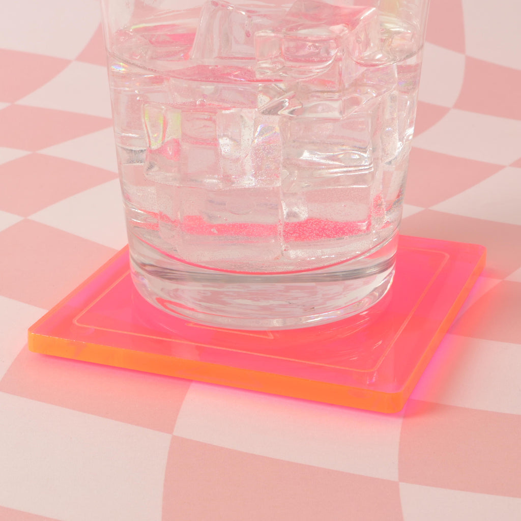 A clear glass filled with ice and a drink, resting on a neon pink square coaster. The background is a pink and white checkered pattern.