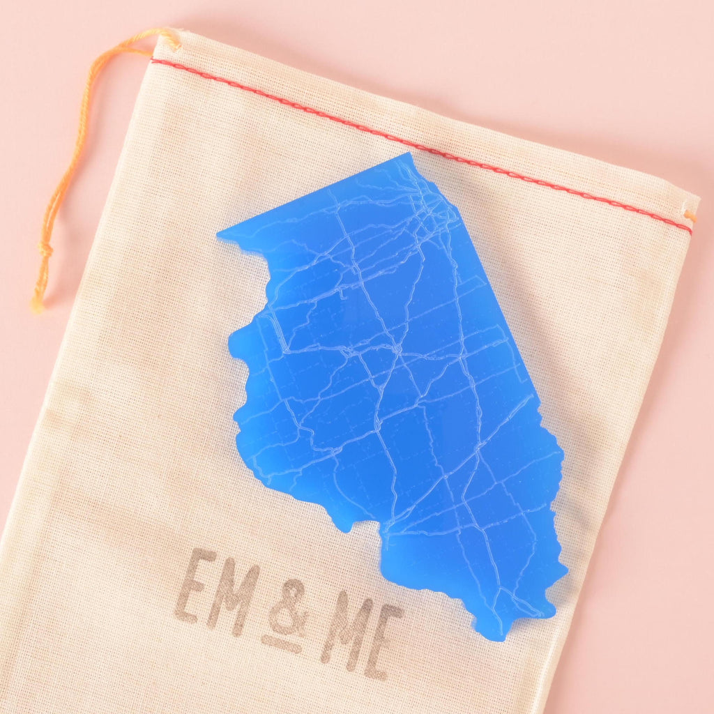Blue acrylic coaster shaped like the state of Illinois with engraved road map, displayed on a beige EM & ME drawstring pouch.
