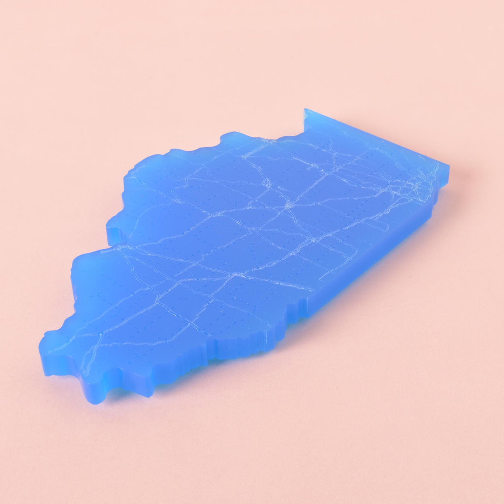 Blue acrylic coaster shaped like the state of Illinois with engraved roadway map design, displayed on a pink background.