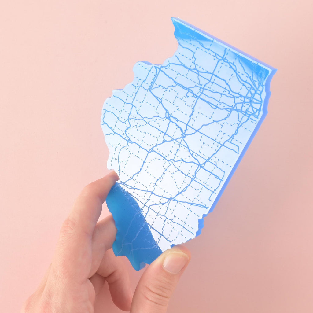Hand holding a blue acrylic coaster shaped like the state of Illinois with engraved roadway design, shown against a pink background.