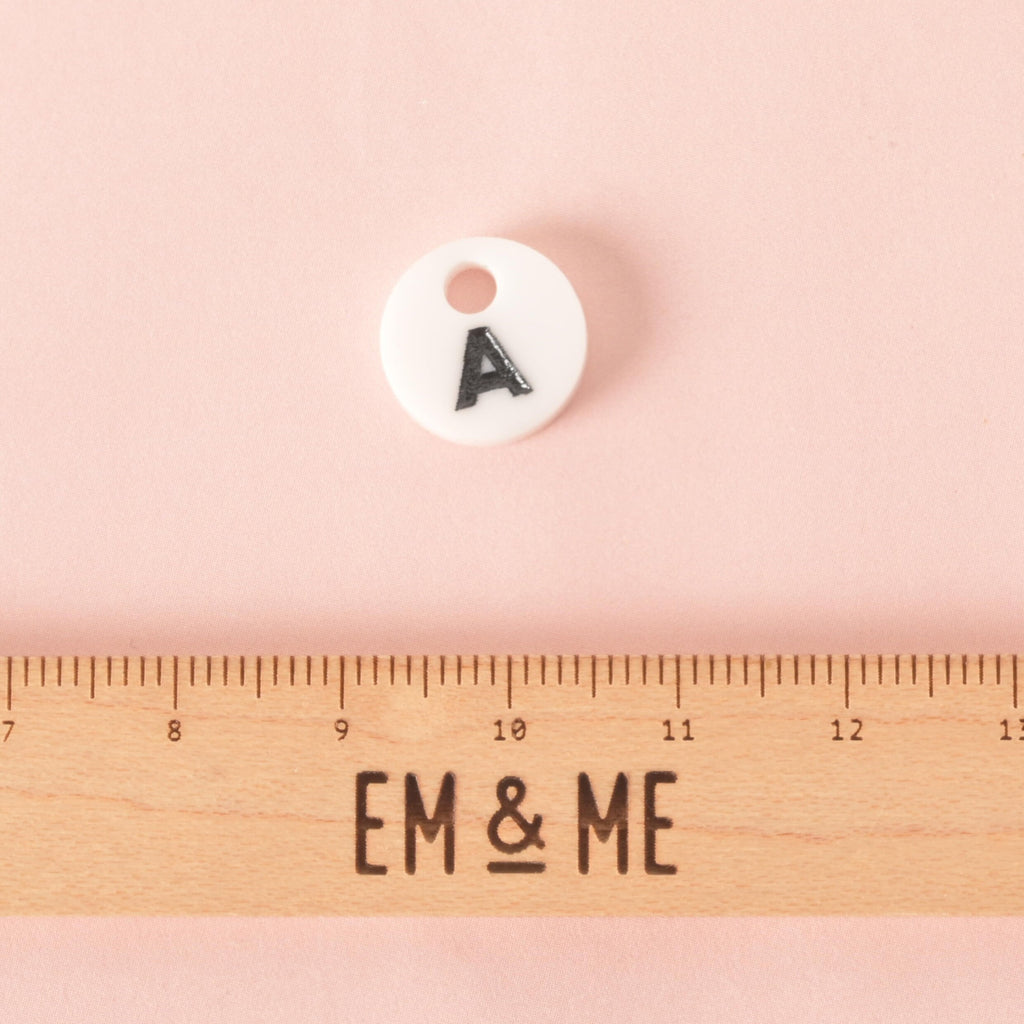 A small, white, circular pet tag charm with a black letter "A" in the center. The charm is displayed above a wooden ruler with the logo "EM & ME." The background is a solid pink.