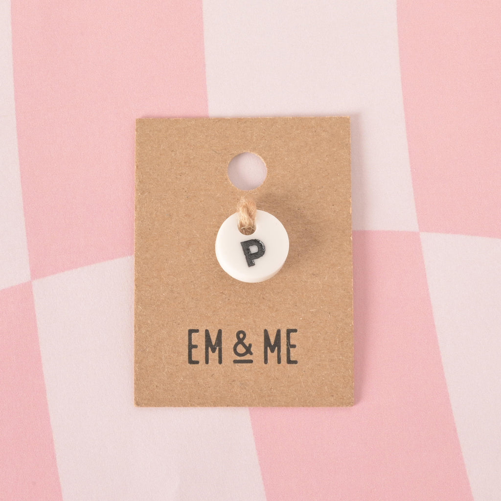A small, white, circular pet tag charm with a black letter "P" in the center. The charm is attached with a piece of string to a brown cardboard tag with the logo "EM & ME." The item is displayed on a pink and white checkered background.