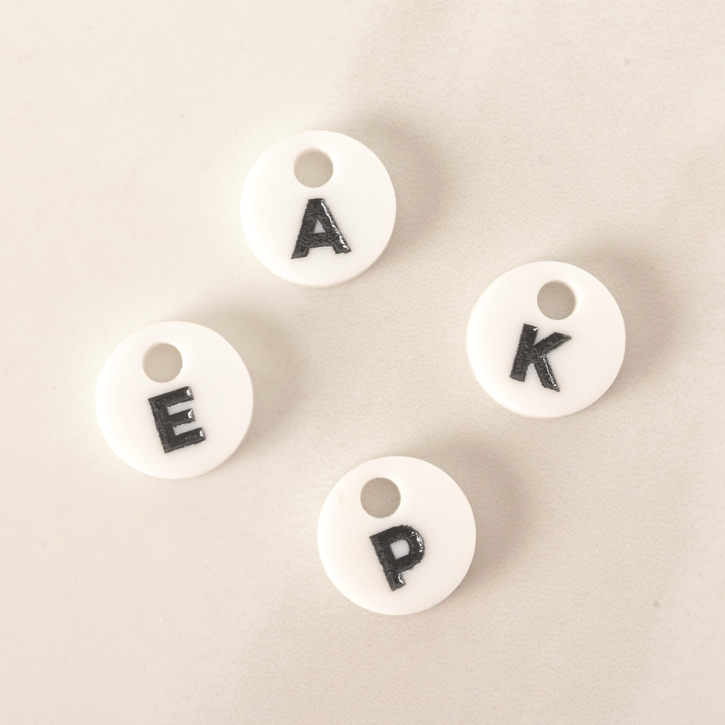 A group of four small, white, circular pet tag charms, each with a single black letter in the center. The visible letters are "A," "E," "P," and "K." The charms are arranged on a light-colored, speckled countertop or stone surface.