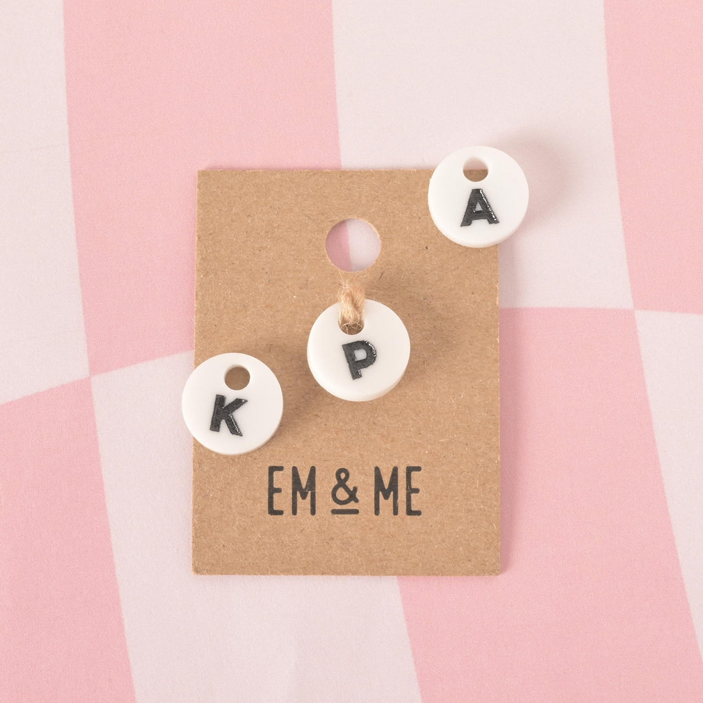 A group of three small, white, circular pet tag charms, each with a single black letter in the center. The visible letters are "K," "P," and "A." The charm with the letter "P" is attached with a string to a brown cardboard tag with the logo "EM & ME." The other two charms are loose beside it. The items are displayed on a pink and white checkered background.