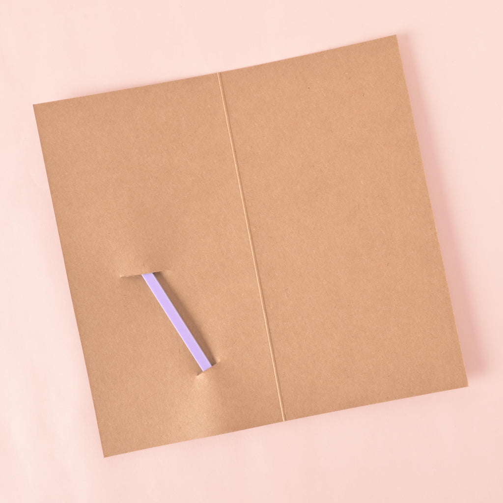 An blank brown gift card, folded in half, with a slit cut into it to hold a stirrer. The back of a light purple cocktail stirrer is partially visible through the slit. The background is a solid pink color.