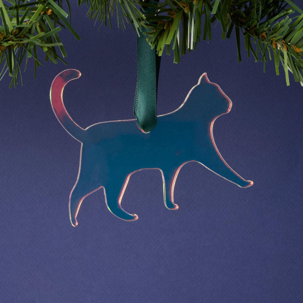 A laser-cut cat silhouette ornament made from iridescent duochrome acrylic (shifting between dark blue and green/pink). The ornament is hanging by a dark green ribbon from a pine branch on a dark background.