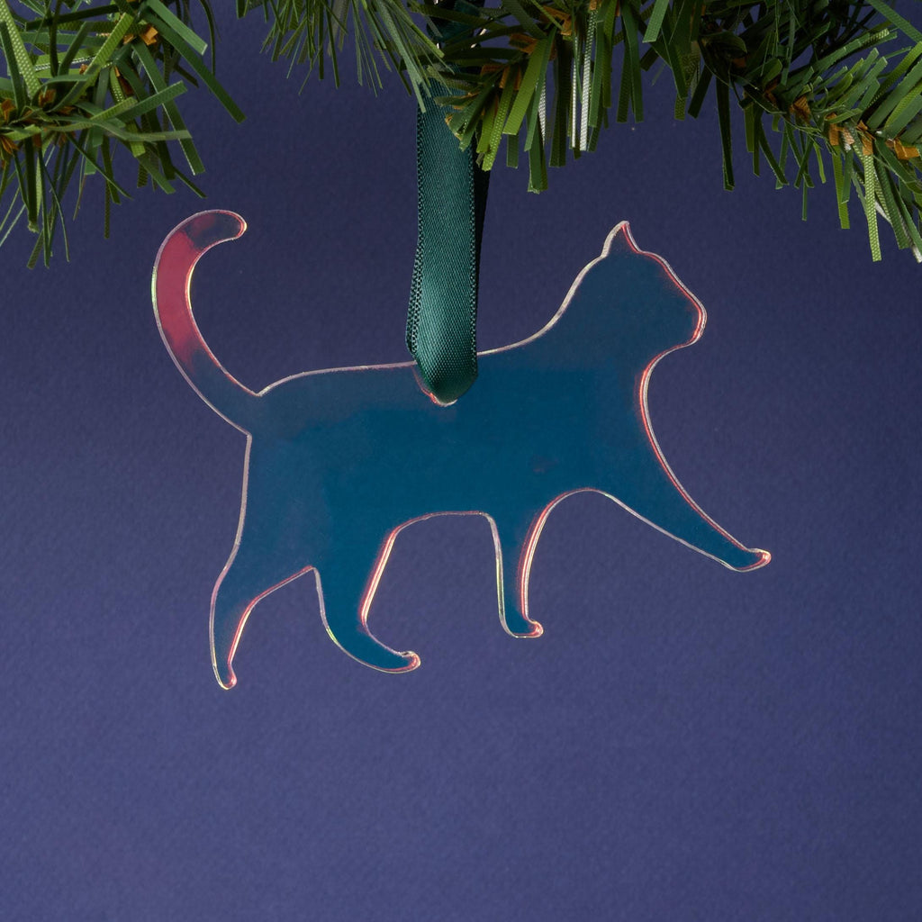 A laser-cut cat silhouette ornament made from iridescent duochrome acrylic (shifting between dark blue, green, and pink). The ornament is hanging by a dark green ribbon from a pine branch on a dark background.