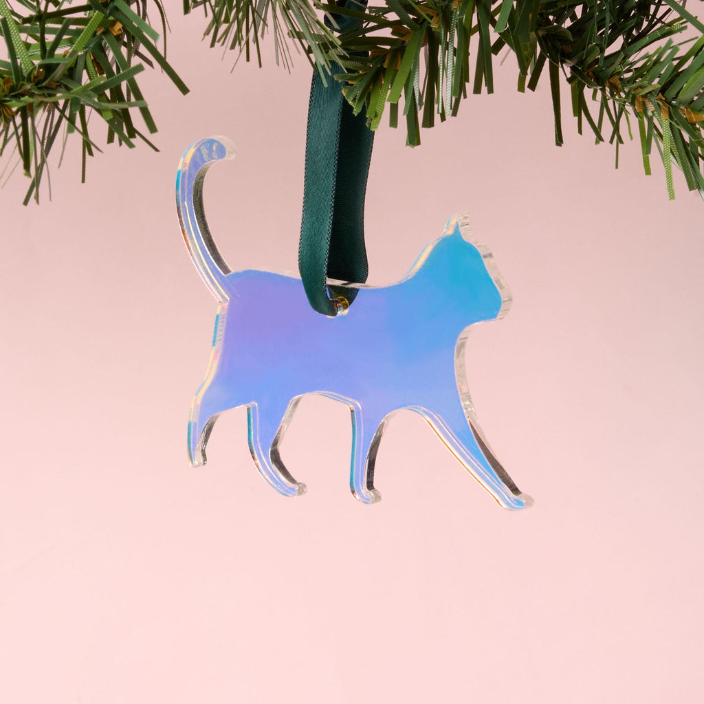 A laser-cut cat silhouette ornament made from iridescent duochrome acrylic, shifting between blue, purple, and pink. The ornament is hanging by a dark green ribbon from a pine branch on a pink background.