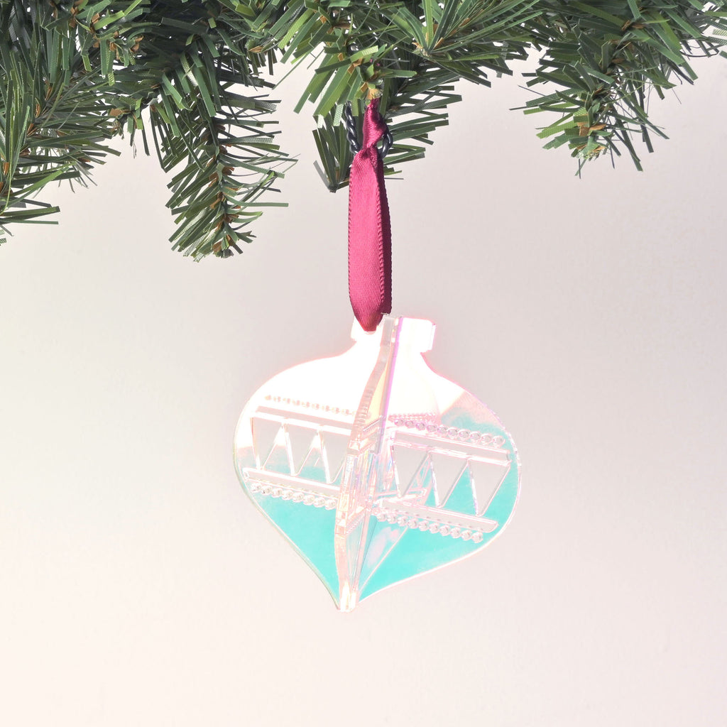 A iridescent, flat-pack Christmas tree ornament shaped like a classic glass ornament. The assembled ornament is hanging from a pine branch with a dark black ribbon. The ornament has a zig-zag and dotted design etched on the sides. The background is a solid white color.