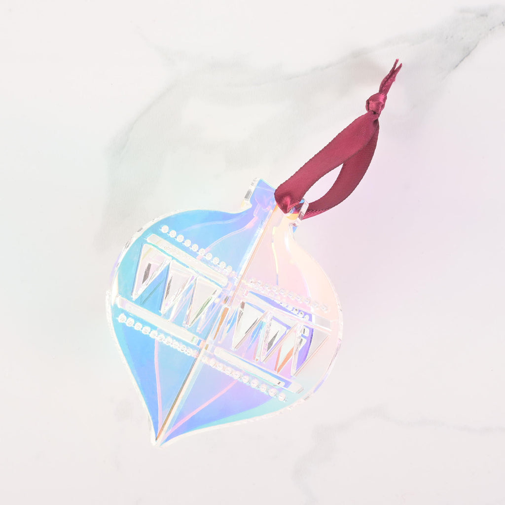 A Iridescent flat-pack Christmas tree ornament shaped like a classic glass icicle ornament. The ornament is assembled and has a dark black ribbon tied to the top for hanging. The item is displayed on a white marble surface.