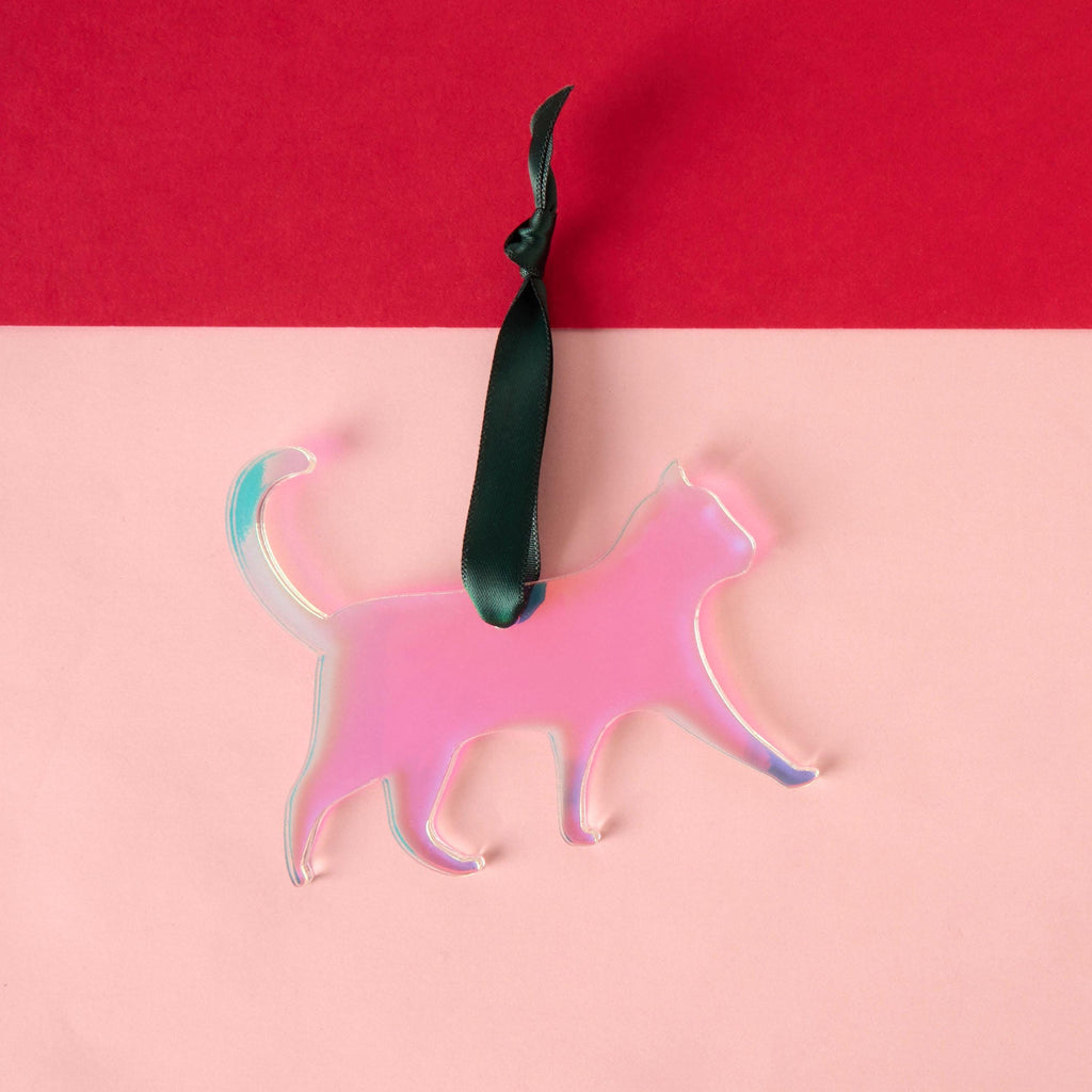 A laser-cut cat silhouette ornament made from vibrant iridescent pink acrylic that shifts colors (duochrome). The ornament is hanging from a dark green satin ribbon and is displayed against a two-tone pink and red background.