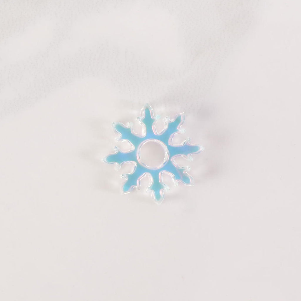 A close-up shot of a iridescent snowflake-shaped charm with glitter accents and a hole in the middle. The charm is displayed on a white marble surface.