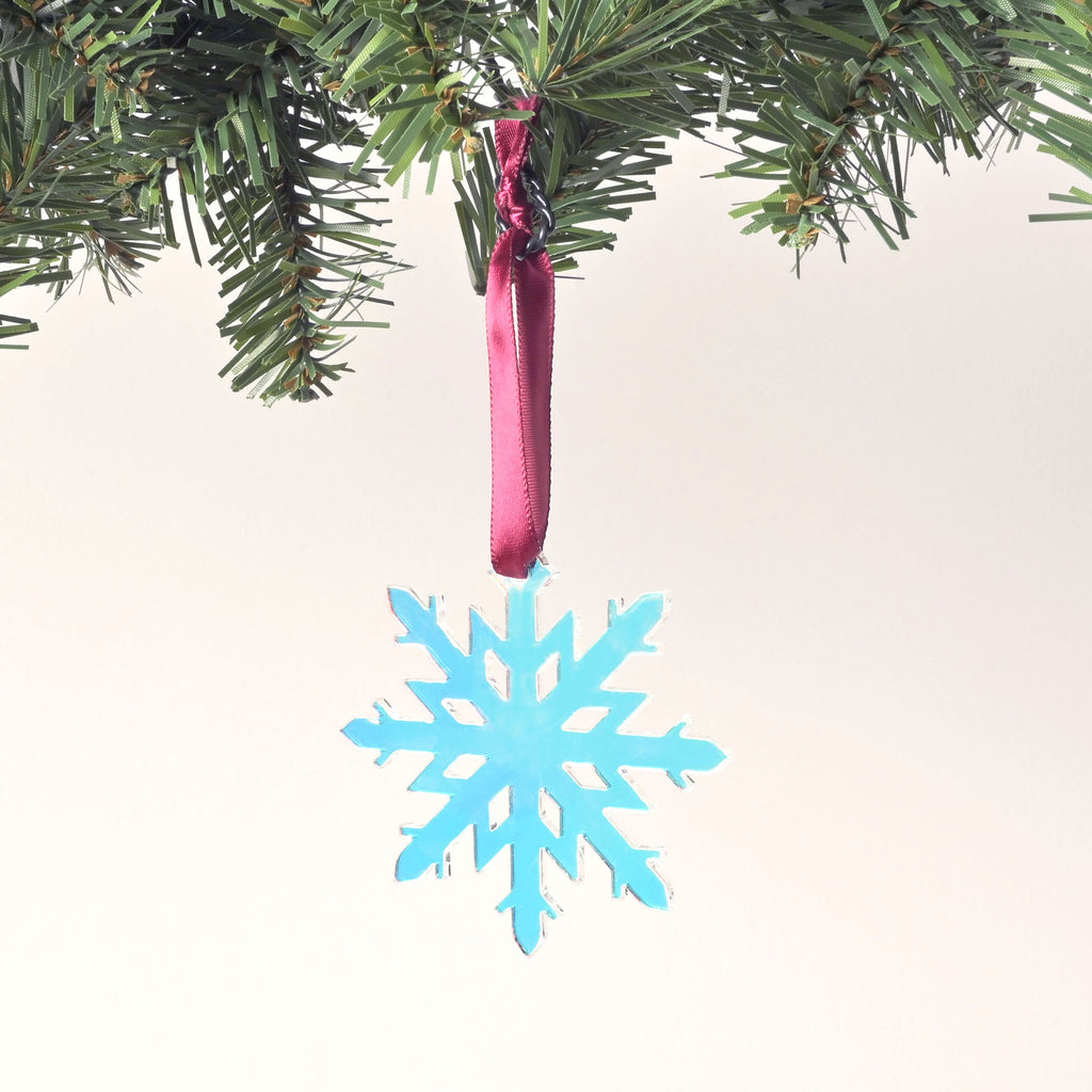 A close-up shot of an iridescent snowflake ornament with a pinkish and bluish sheen. A dark red ribbon is tied to the top, and the ornament is hanging from a pine branch against a solid white background.