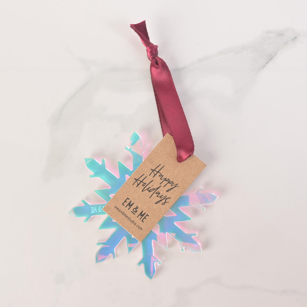 A close-up shot of an iridescent snowflake ornament with a pinkish and bluish sheen. A brown cardstock tag that says "Happy Holidays" and "EM & ME" is attached to the ornament with a dark red ribbon. The item is displayed on a white marble surface.