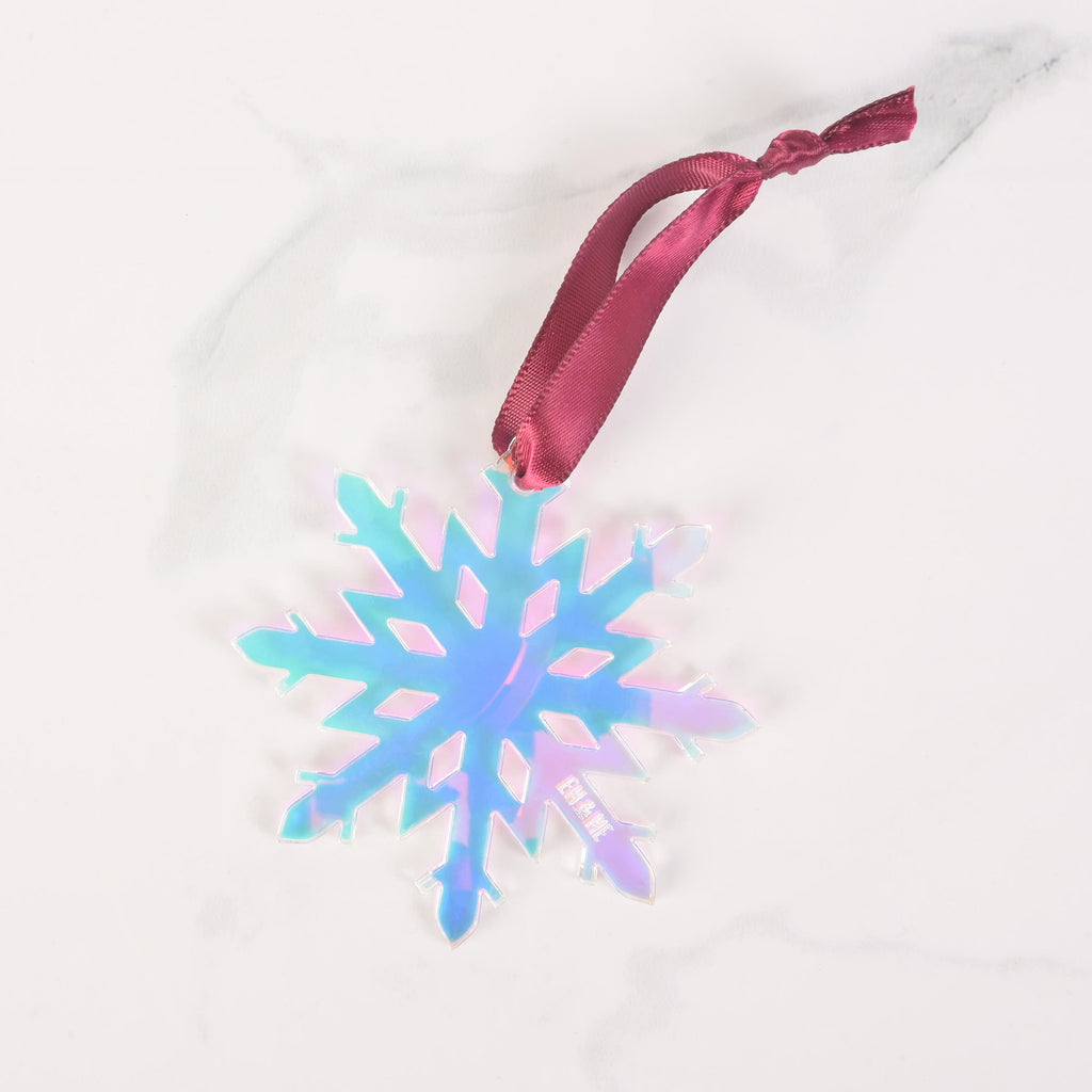 A close-up shot of an iridescent snowflake ornament with a pinkish and bluish sheen. A dark red ribbon is tied to the top for hanging. The ornament is displayed on a white marble surface and has the "EM & ME" logo faintly etched on the bottom right.
